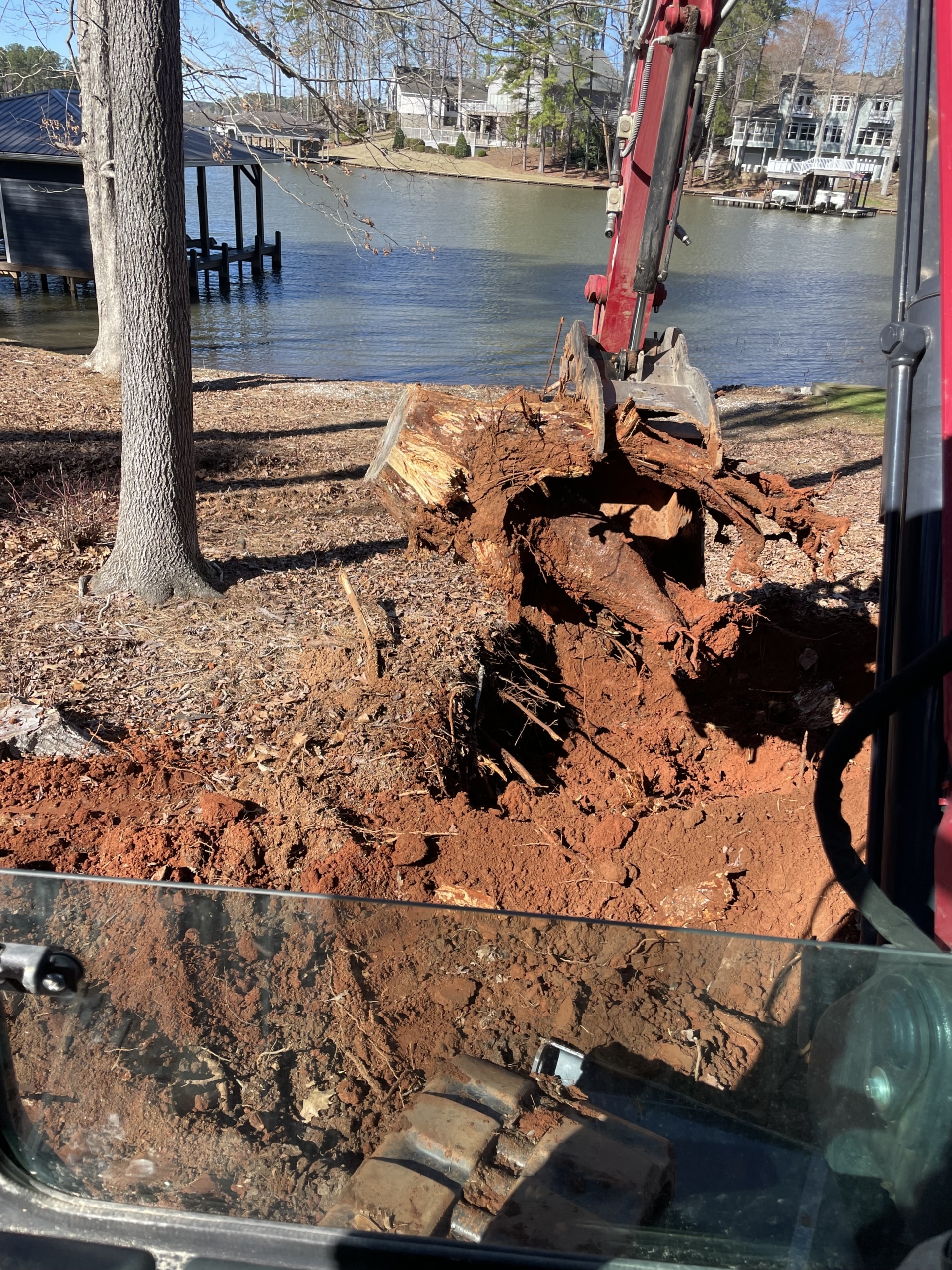 Excavator digging into the ground near a body of water, showing exposed soil and tree roots.