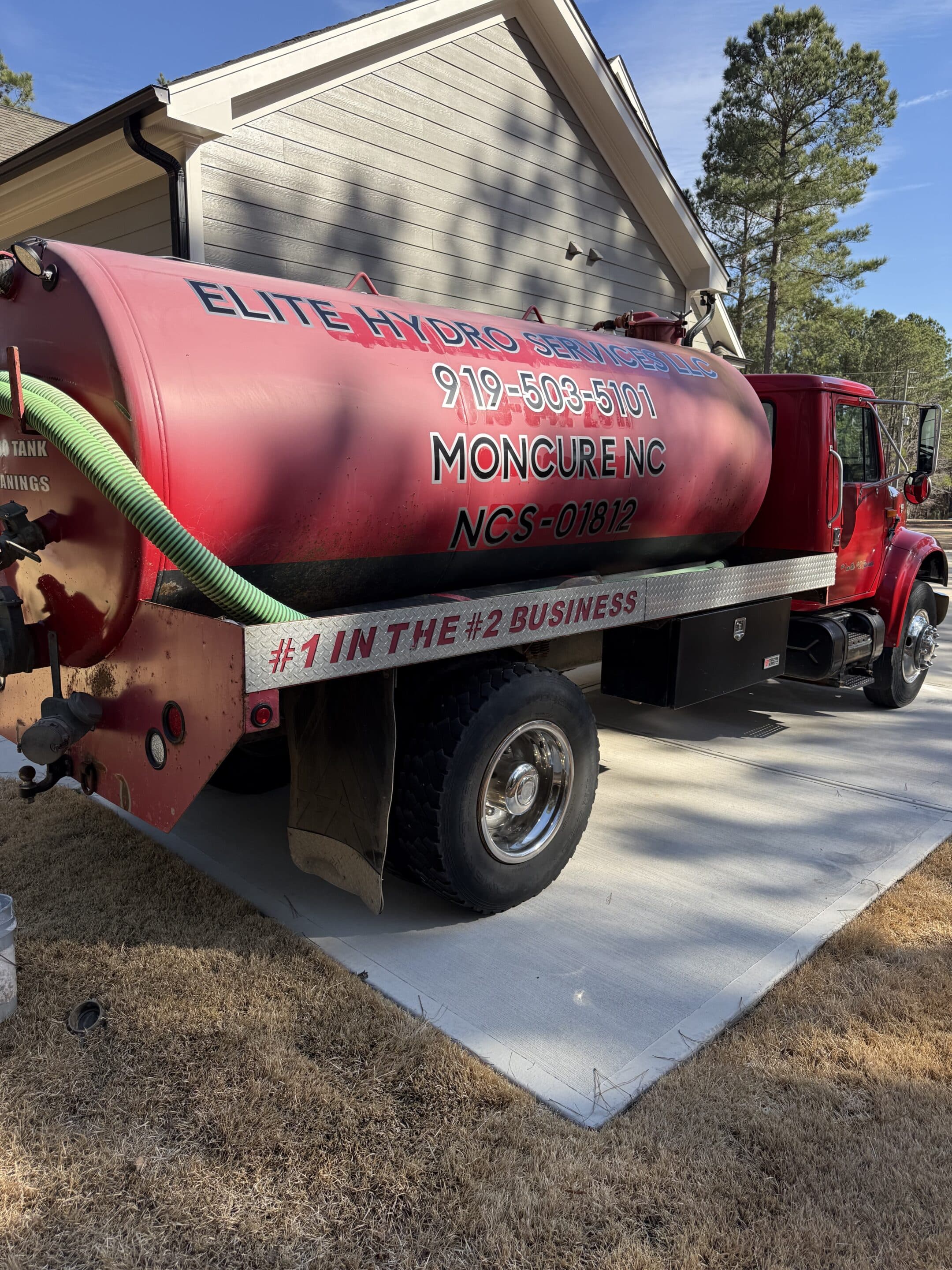 Red Elite Hydro Services septic truck with green hose, parked beside a house in Moncure, NC. The vehicle displays contact info and the slogan "#1 in the #2 Business."